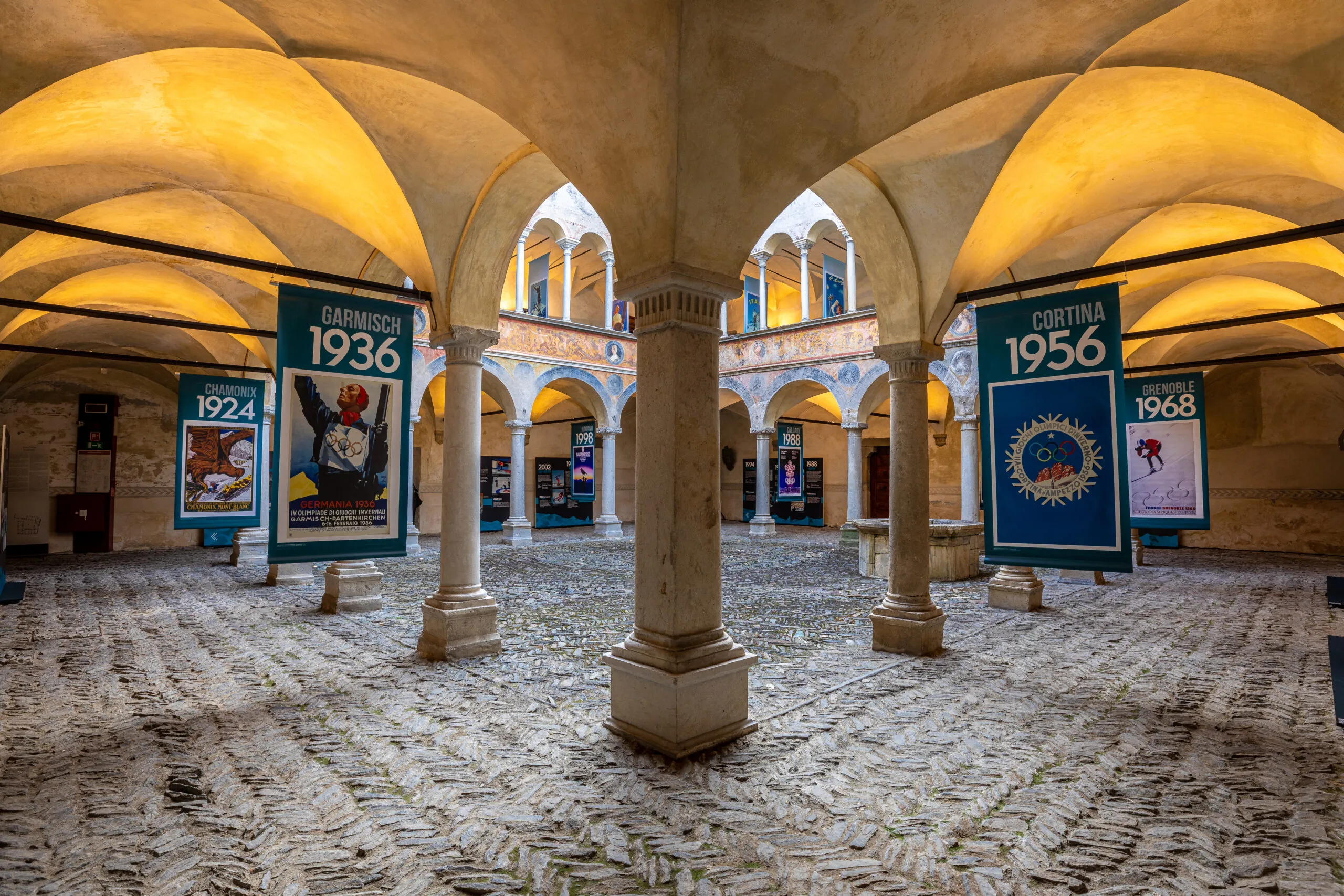 Appesi come stendardi sotto ai portici del loggiato nella corte, i manifesti delle passate edizioni olimpiche scandiscono la timeline delle Olimpiadi e Paralimpiadi invernali.